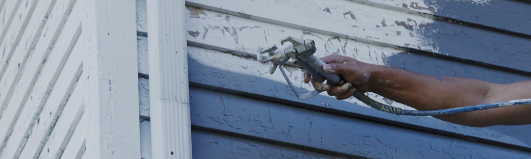 Person painting a house exterior with a paint sprayer.
